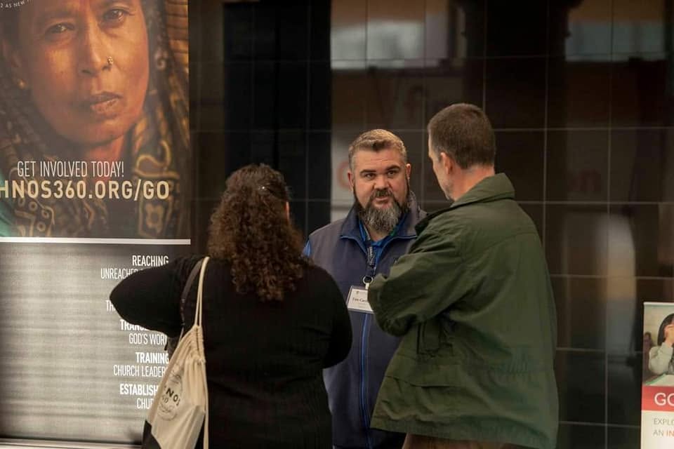Tim Carmical speaking one-on-one with a visitor beside an Ethnos360 display